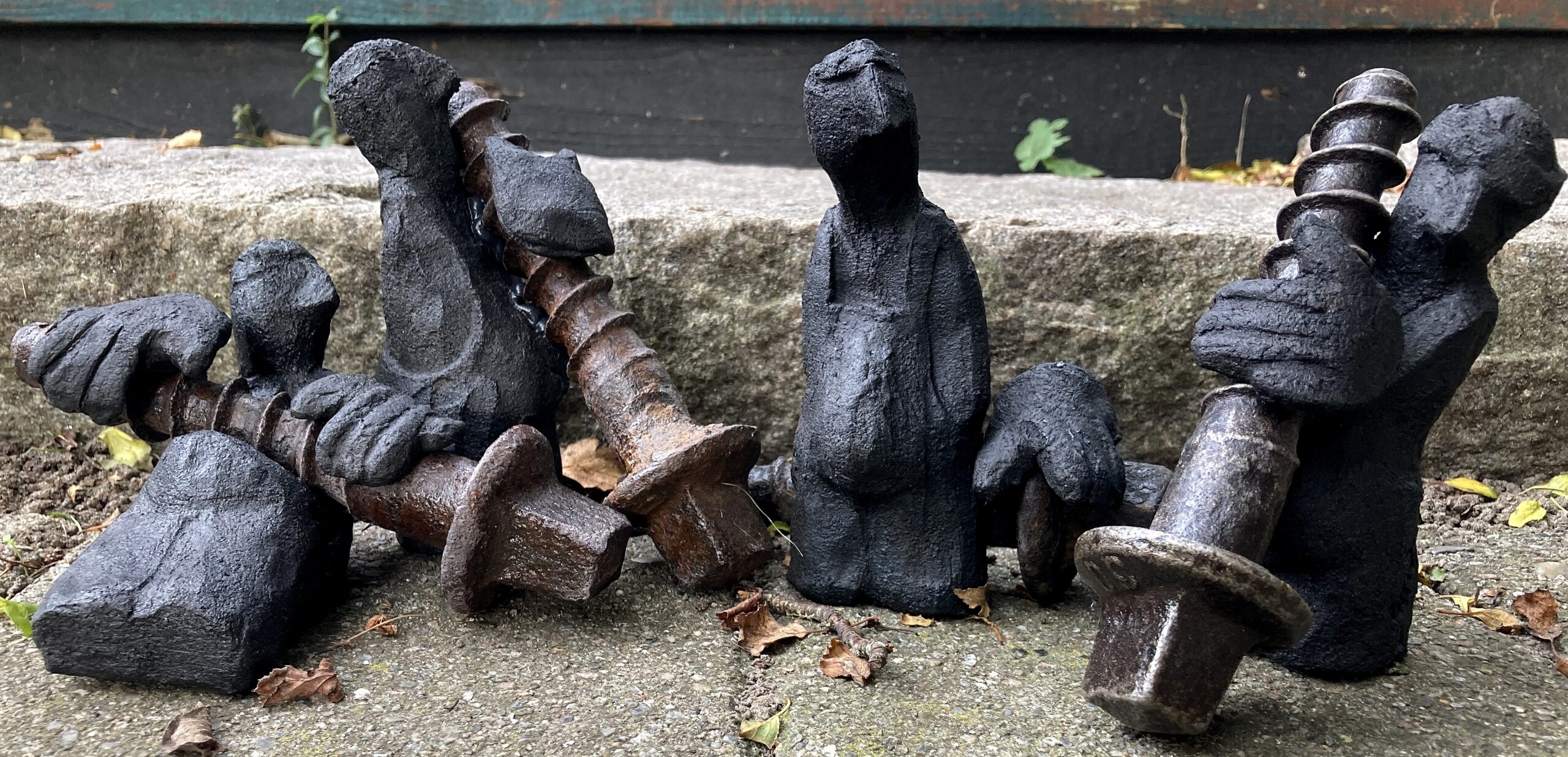 Fem abstrakte, mørkfarvede menneskelignende skulpturer er placeret på et stentrin, og nogle af dem holder store rustne bolte. Nedfaldne blade og affald ligger spredt rundt omkring.